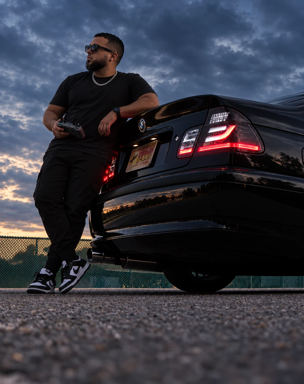 Jonathan Huskey leaning on his black BMW E46 at dusk, drone controller in hand