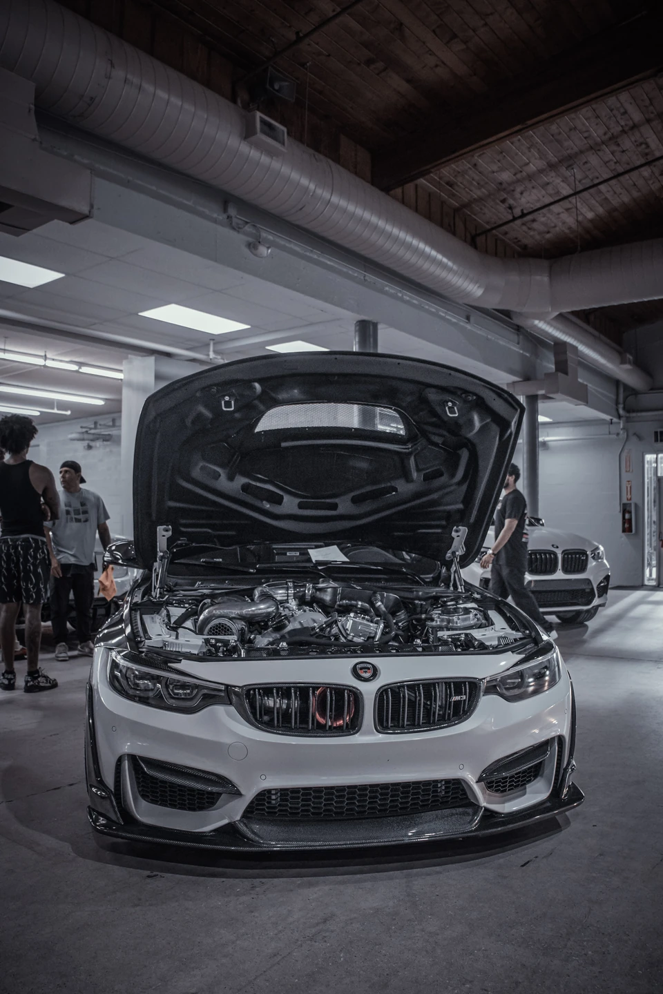 White BMW M coupe with its hood up in a cool-toned industrial warehouse