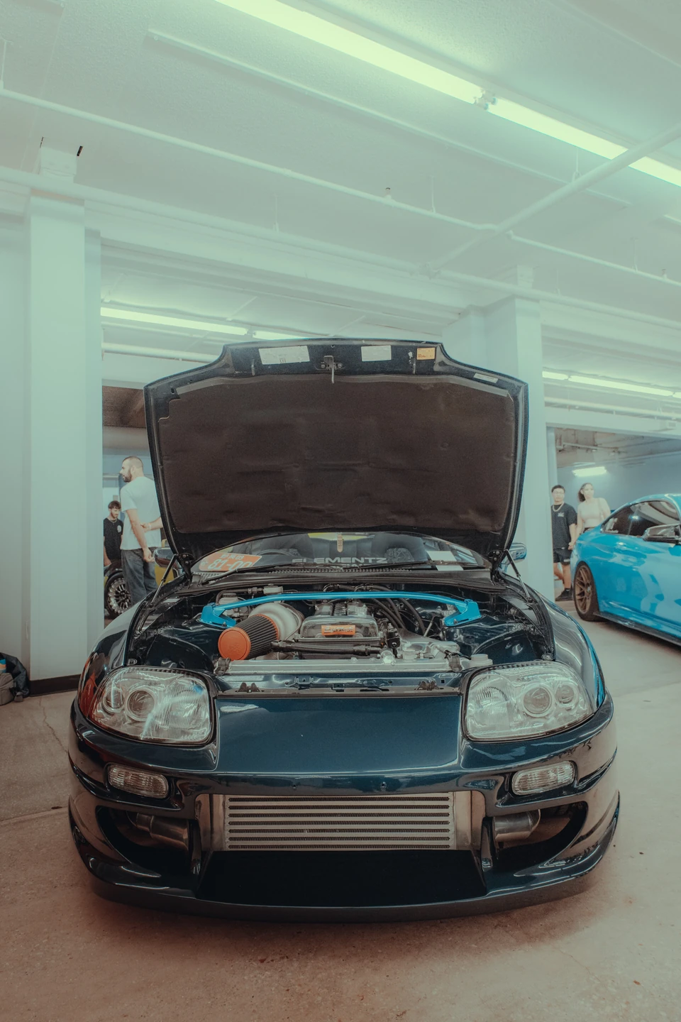 Dark teal Toyota Supra with its hood raised under fluorescent lights, in a faded film-style grade