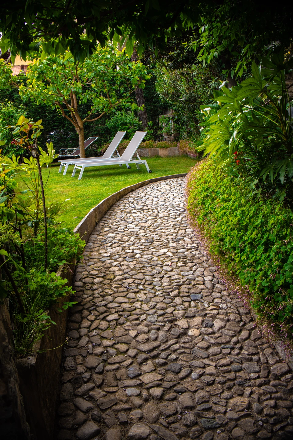 Curving cobblestone path through a lush green garden with white lounge chairs