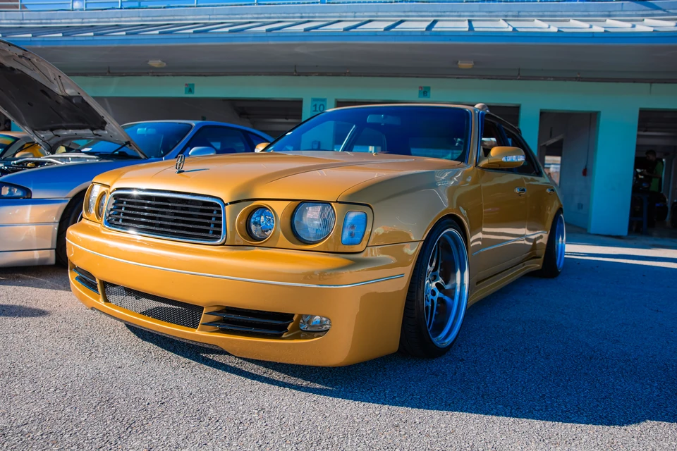 Mustard-yellow luxury sedan on polished deep-dish wheels parked in front of numbered pit garages
