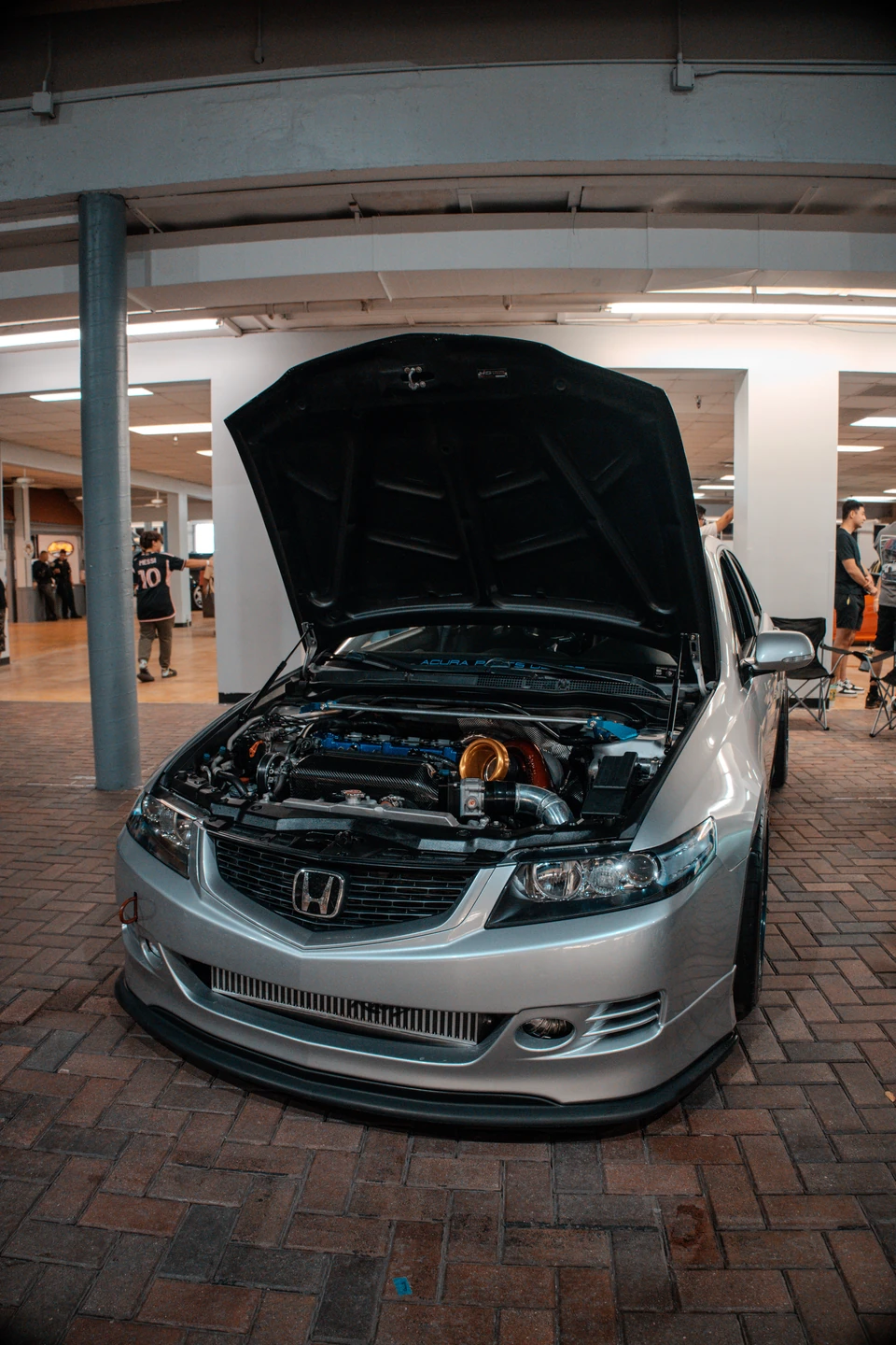 Lowered silver Honda sedan with its hood raised on a brick floor at an indoor car show