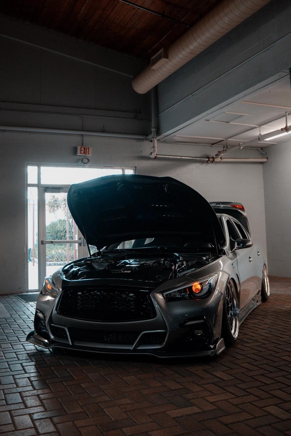 Dark gray Infiniti sedan with its hood open in a dim warehouse under a wood ceiling