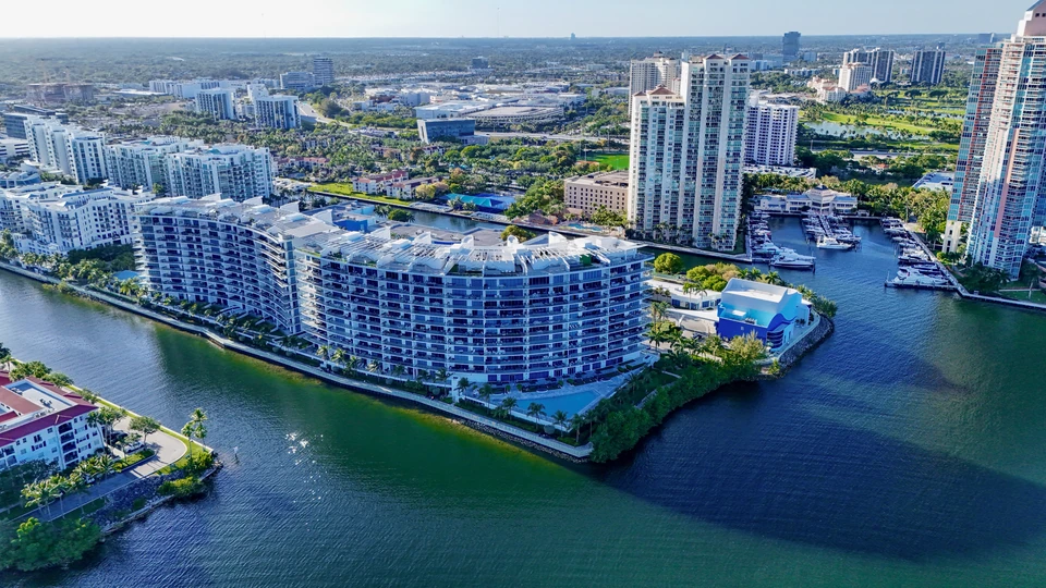 Aerial view of a curved waterfront condo building and marina along a wide blue waterway