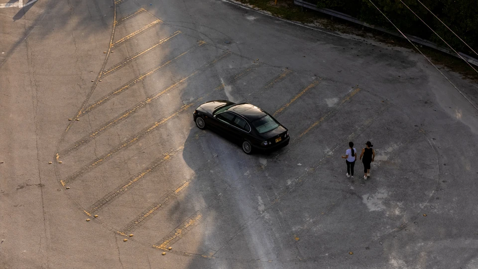 Aerial view of a black BMW sedan on a weathered lot in late light, with two people walking toward it
