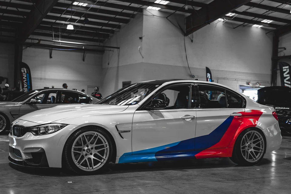 Black and white photo of a white BMW sedan in which only its blue and red M stripes keep their color