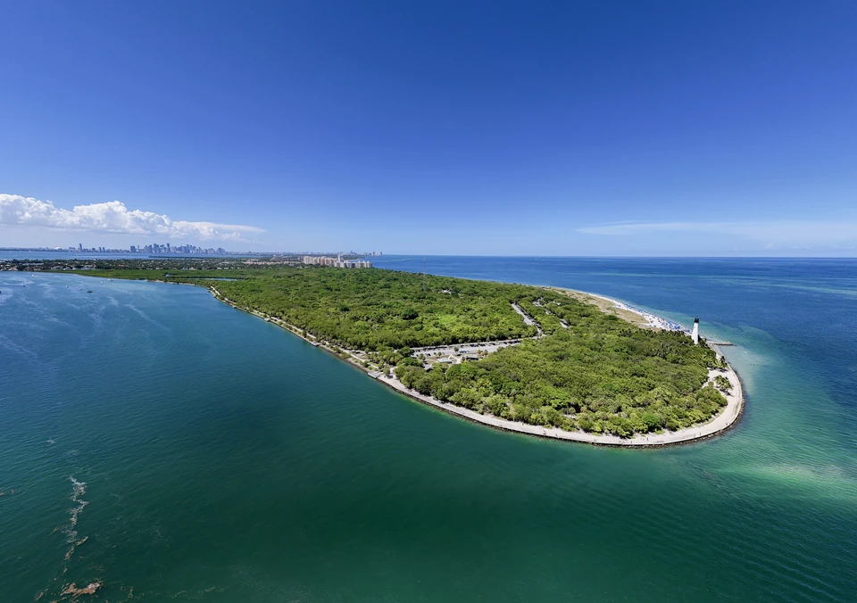 Aerial view of a green peninsula curving into turquoise water with the Miami skyline on the horizon
