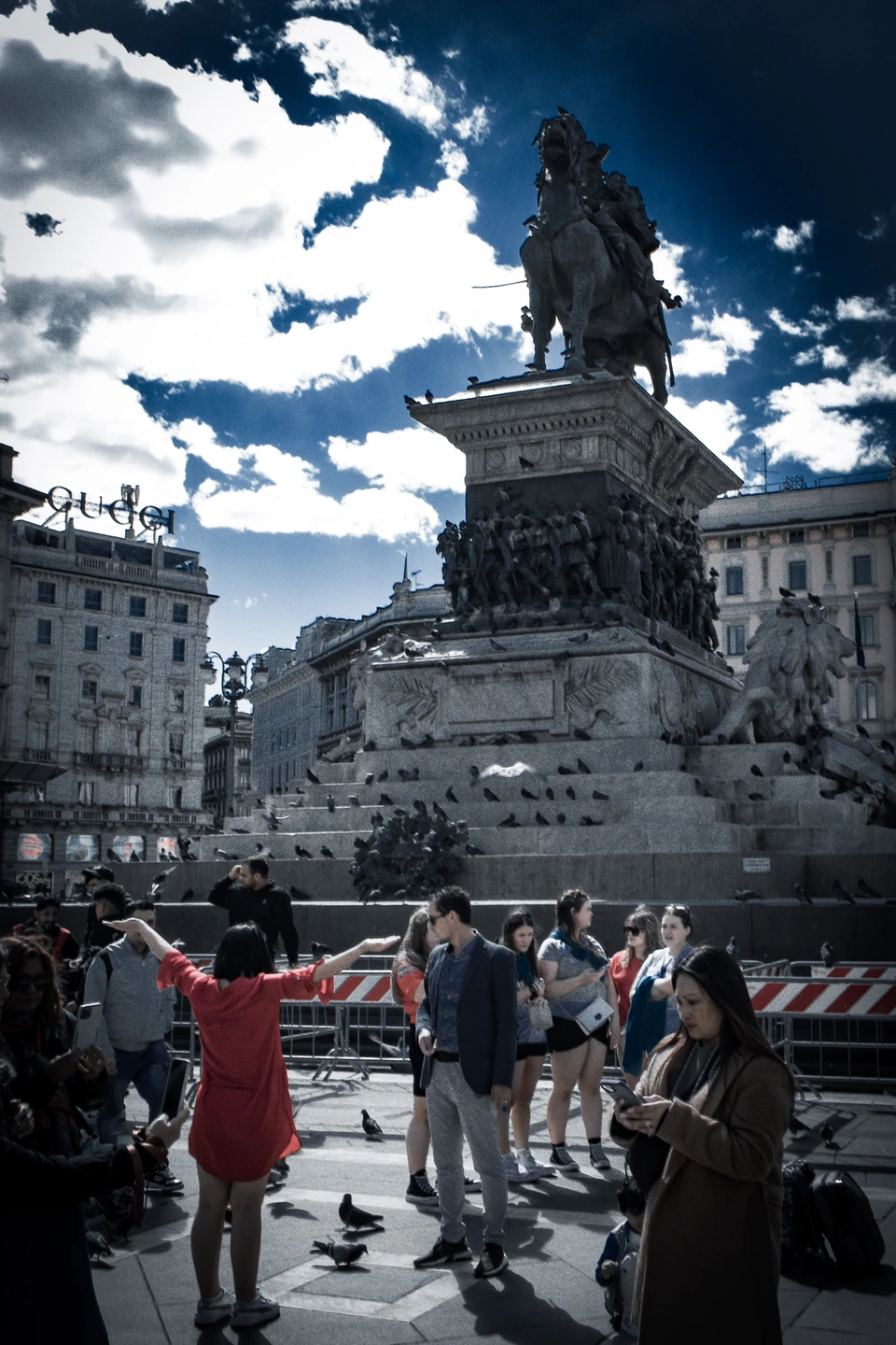 Equestrian monument in Piazza del Duomo under a dramatic sky, with crowds and pigeons in the square
