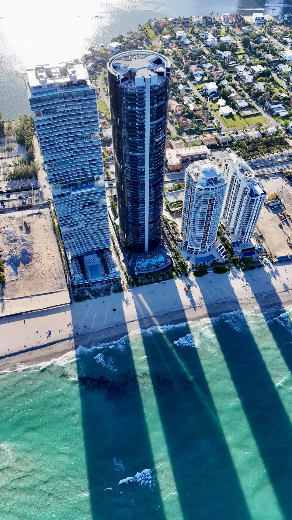 Aerial view looking down on beachfront towers whose shadows stretch across the sand into turquoise water