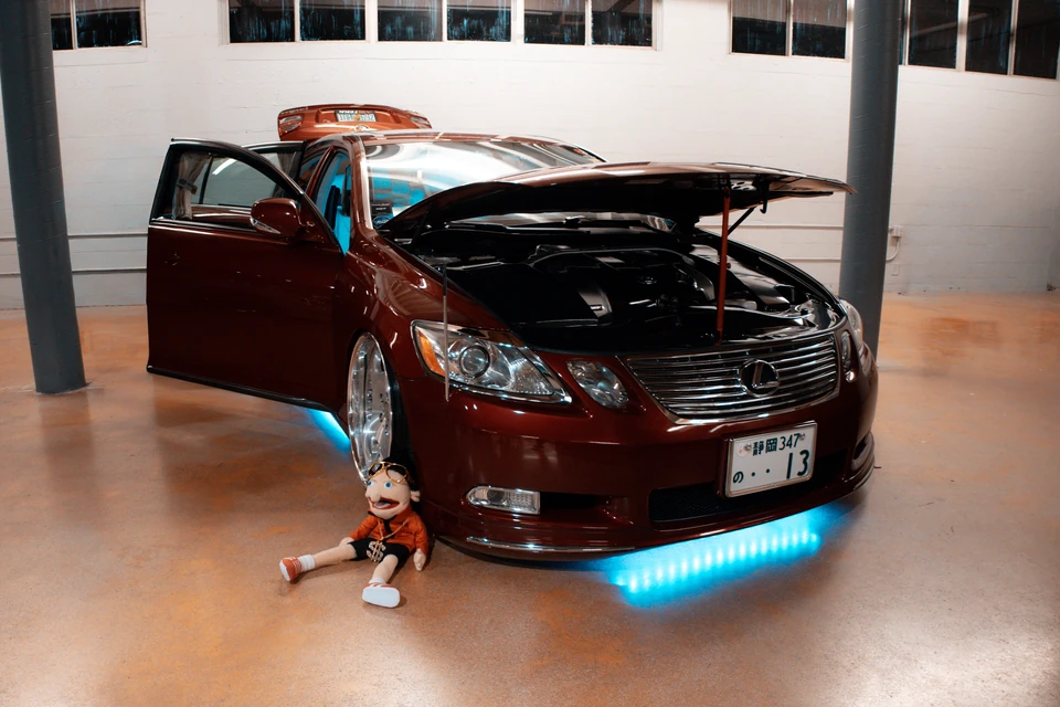 Lowered maroon Lexus sedan with blue underglow and a plush doll sitting by its front bumper