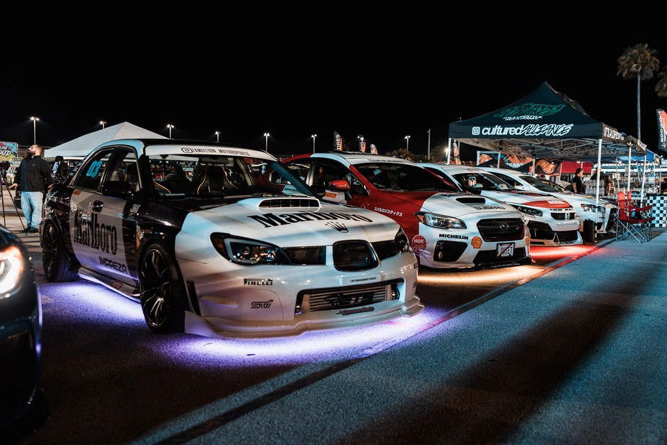 Row of rally-livery Subaru sedans lit by colored underglow at a night car meet