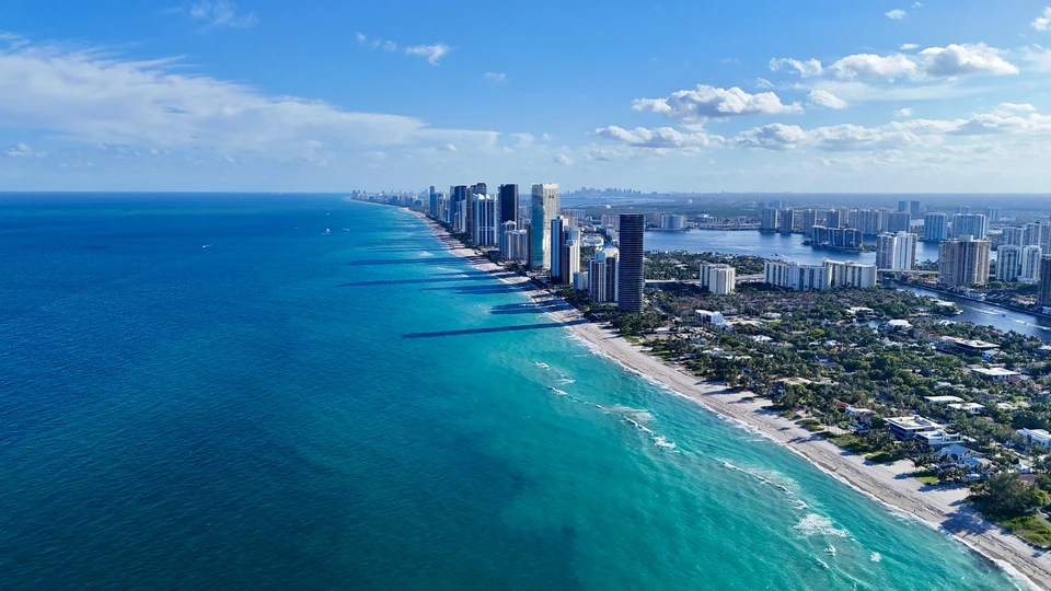 Aerial view down a long beach lined with high-rise towers, turquoise water and scattered clouds
