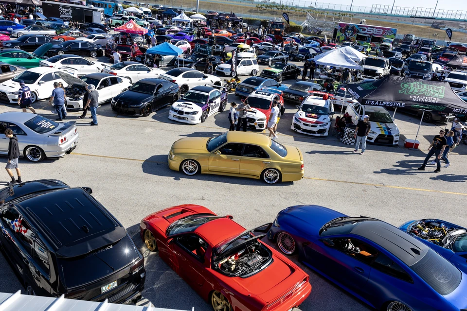 Elevated view over a packed car meet in a pit lane, with rows of tuned cars and pop-up tents